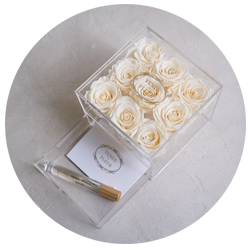 Clear box containing nine cream-colored roses with a gold-accented pen and a card inside the box on a light surface.