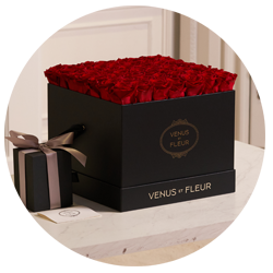 Black square box filled with red roses labeled 'VENUS ET FLEUR' next to a smaller black gift box tied with a brown ribbon, placed on a white surface.