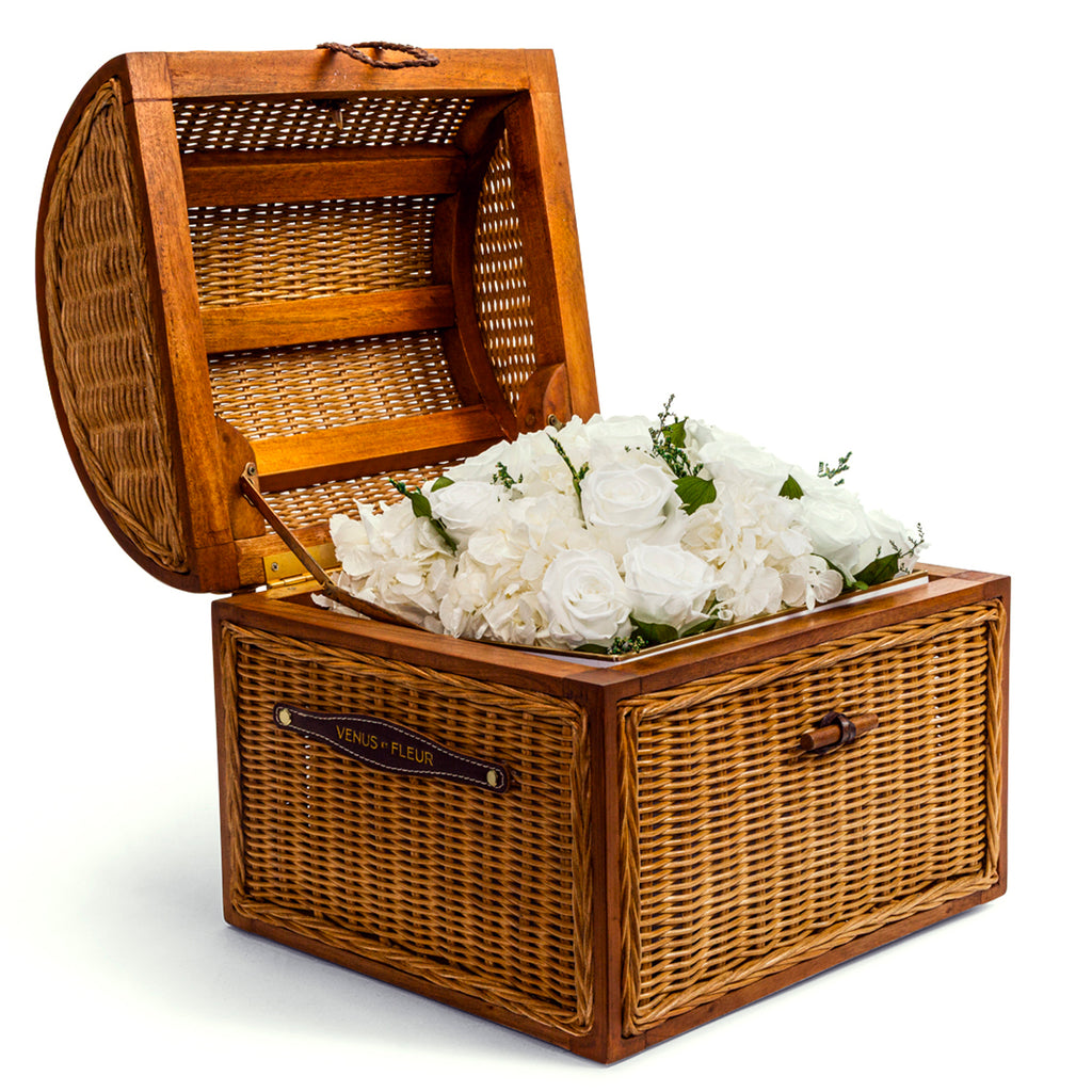Wicker box with open lid containing white roses and hydrangeas, with a leather label reading 'VENUS ET FLEUR'