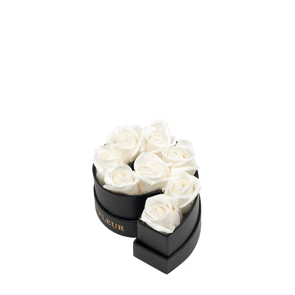 Black heart-shaped box with eight white roses arranged inside, with the word 'FLEUR' visible on the box.
